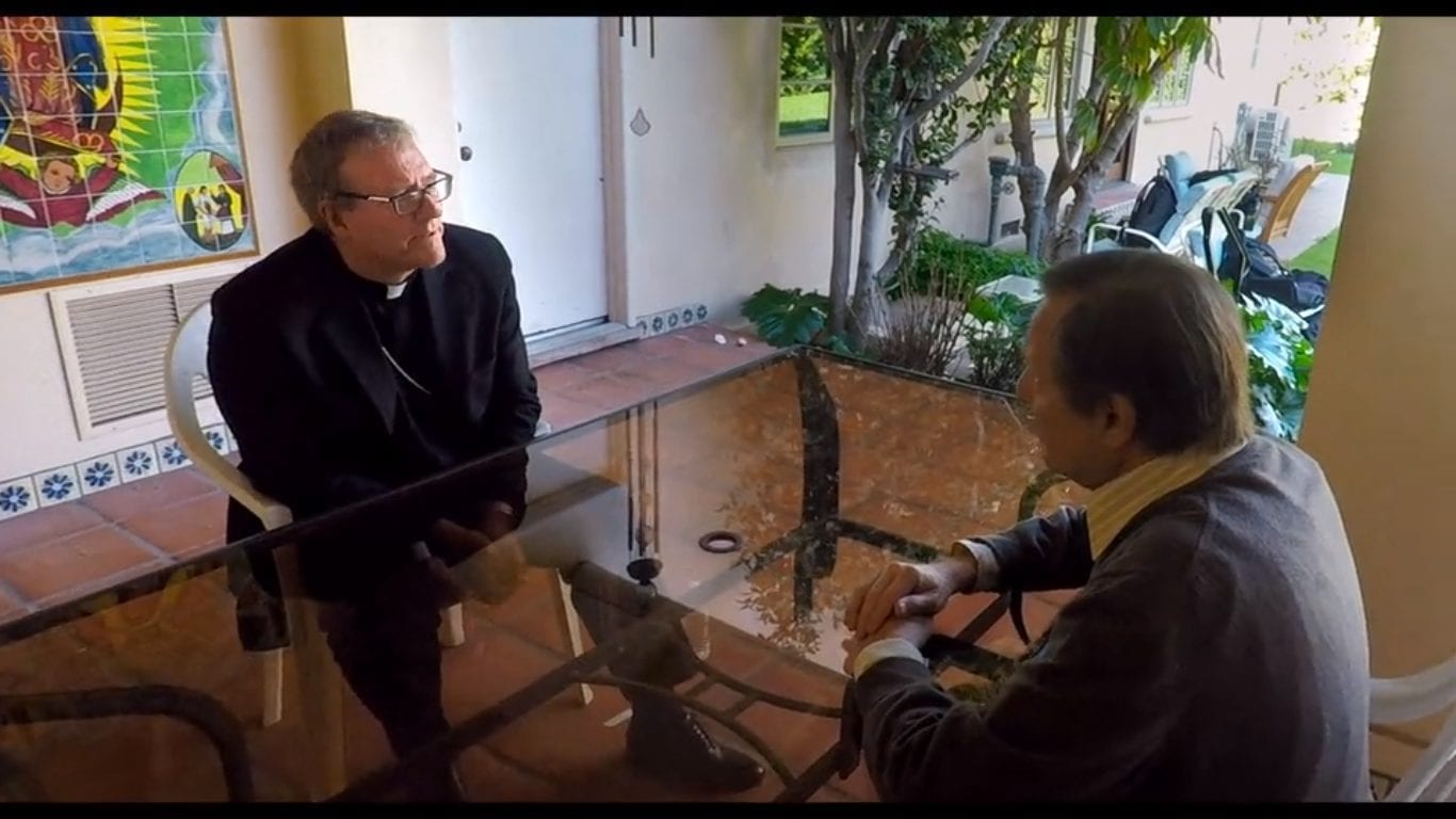 A priest talks to Father Amorth over a glass table