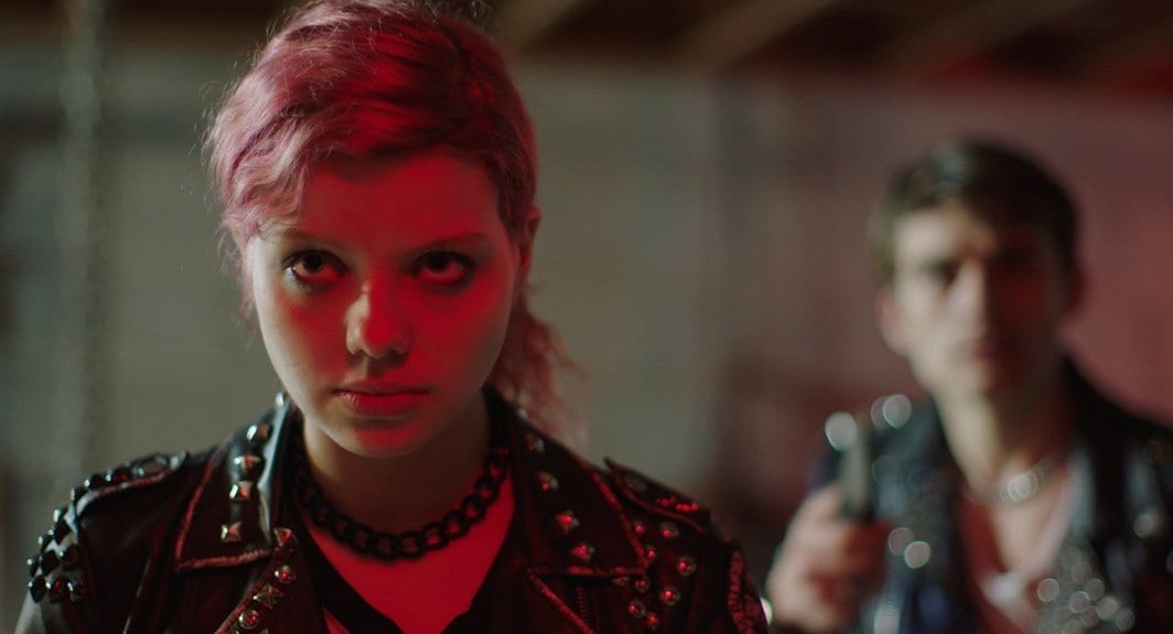 A young woman with pink hair in a studded jacket stands before an out of focus man.