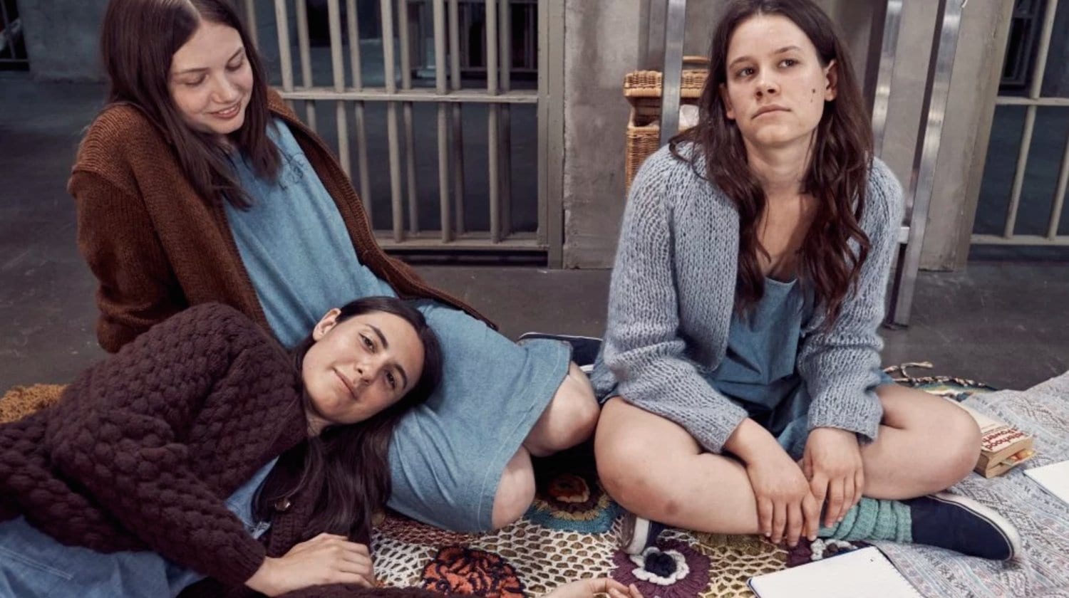 Three girls sit on a blanket, one with her head in another's lap.