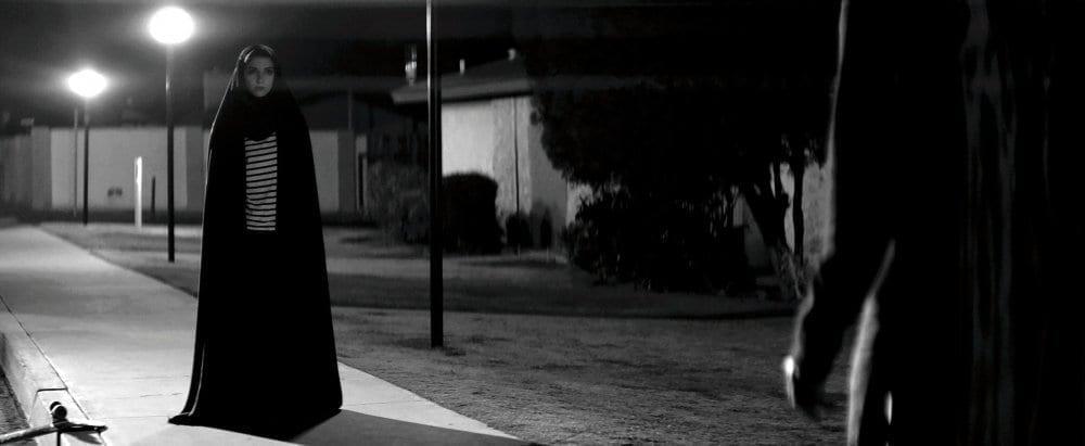 A young girl in a black hooded cloak stands on a dark sidewalk.