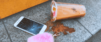 An iphone with a smashed screen, a pink fluffy keyring and a spilled orange and red drink on a step