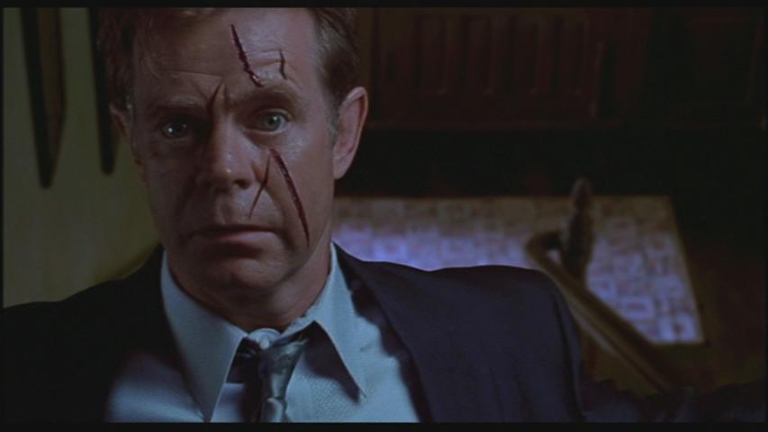 William H. Macy as Detective Arbogast stands at the top of a staircase with a slashed face