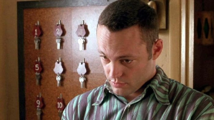 Vince Vaughn as Norman Bates wearing a striped shirt and standing in front of a wall with motel room keys tacked to a board.