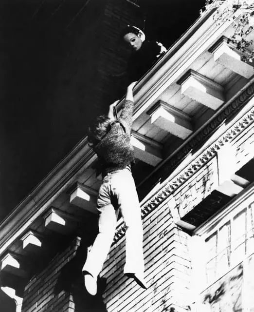 A teenage girl tightly grips a house's gutter, while a man in a mask stands above her on the roof.