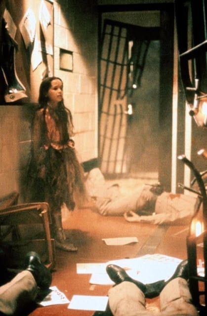 A young girl in a worn out princess costume stands in the middle of a bloodbath at a police station.