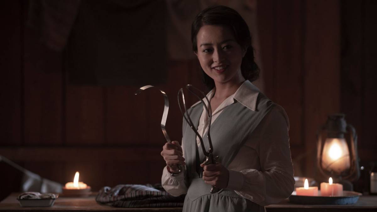 Yuko stands next to a table while brandishing some horrific looking medical equipment