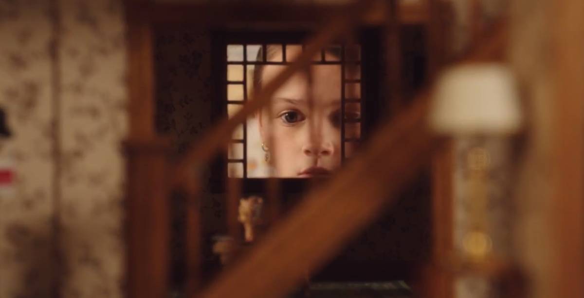 Evie looks into the window of her dollhouse as the severed doll head sits on a table in the foreground.