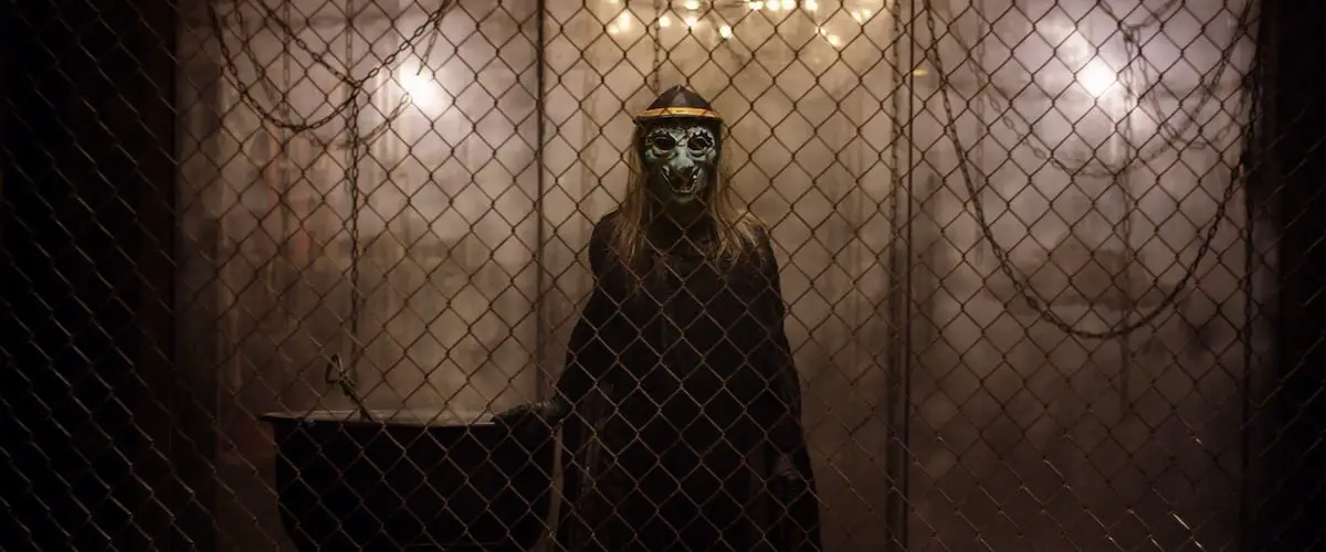 a figure in a witch mask stands behind a wire fence