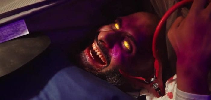 A man contorted in a suitcase with bloodstained teeth and jumper cables plugged to his arm laughs as his eyes glow gold.