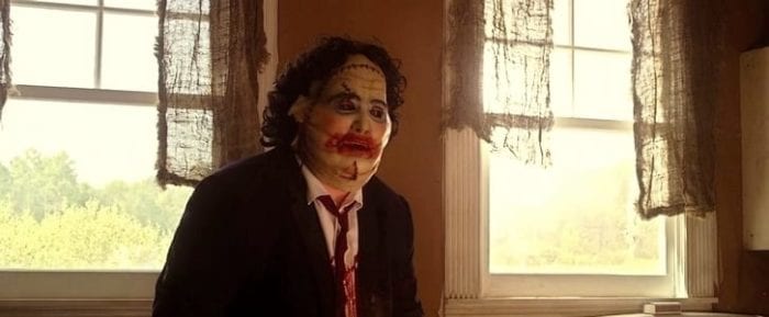 Leatherface sits in a chair in his suit and female mask on