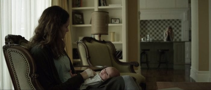 Mary sits on the couch and holds the baby as she stares at Rachel in the kitchen in Still/Born
