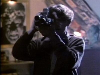 A man looks through a camera with a werewolf poster on the wall behind him.