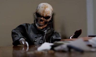 Evil doll, Skull Blade, leans over a table and puts his hook hand on it. 