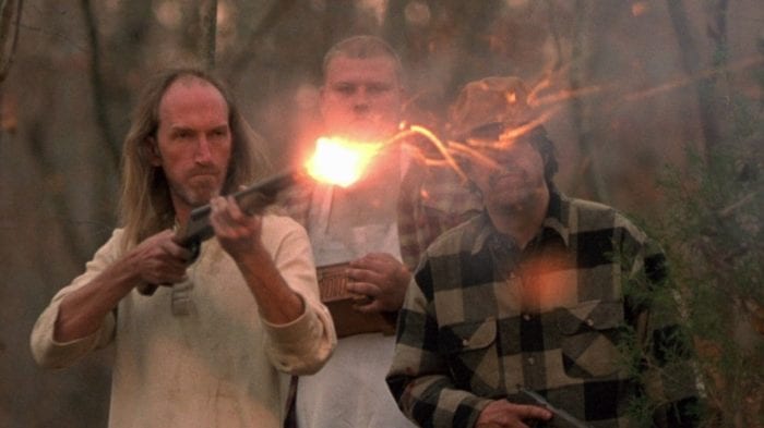 Three local men stand, each armed, while one, a balding man with long hair, fires a double barreled shotgun.