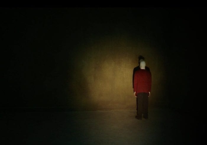 Pretzel Jack, an albino person with a red sweater and brown pants, stands in a corner of a dark room, illuminated only by a flashlight