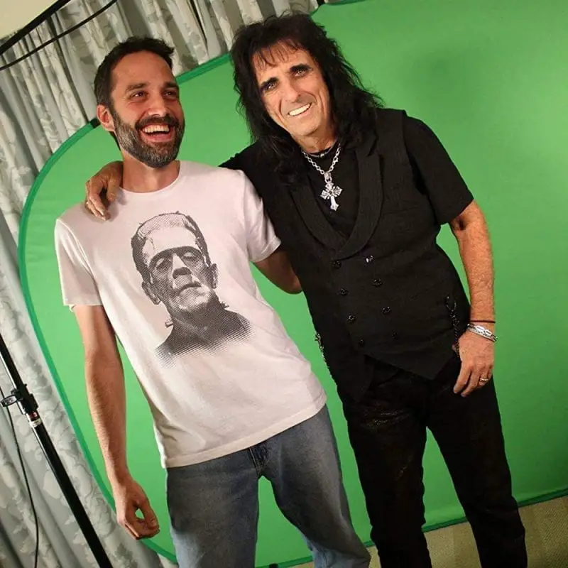 Justin Beahm wears a T-shirt with Frankenstein's monster on it and stands next to Alice Cooper, wearing all black and a cross necklace, in front of a green screen on a production set.
