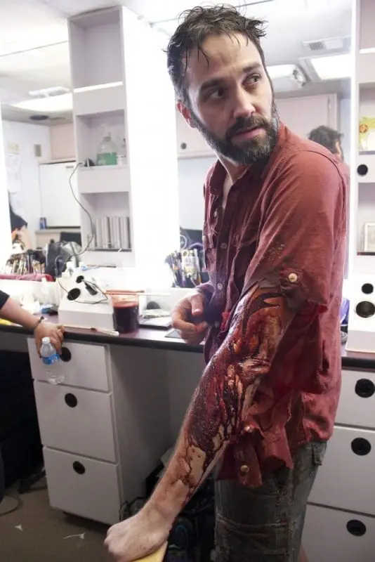 A man is in a special makeup effects studio, with wet hair and a fake wound and blood on his arm.