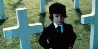 Young Damien standing next to uniform white crosses in a cemetery.