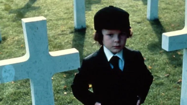 Young Damien standing next to uniform white crosses in a cemetery.