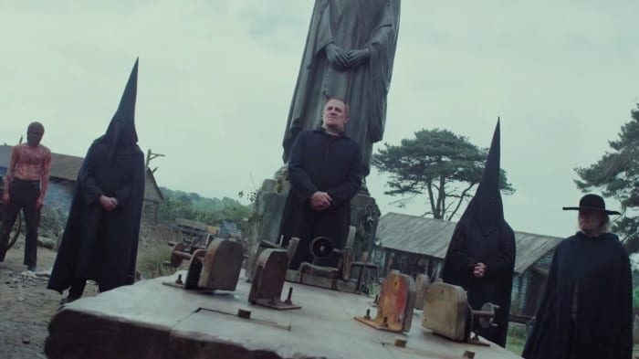 Five figures stand around a block with antiquated machinery in front of a monument. One man is shirtless and bloody, the other men wear all black and two are hooded.