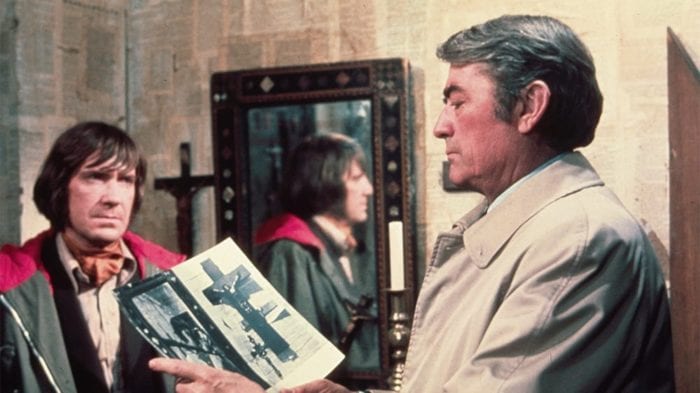 In a room wallpapered with bible pages, Keith Jennings looks on as Robert Thorn studies two photographs.