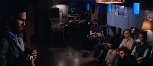 MacReady stands across the room heating the wire to use for blood testing, as the other men sit tied to the couch on the other side of the room.