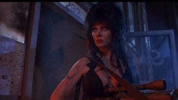 Elvira stands alone holding a machine gun outside, at night.