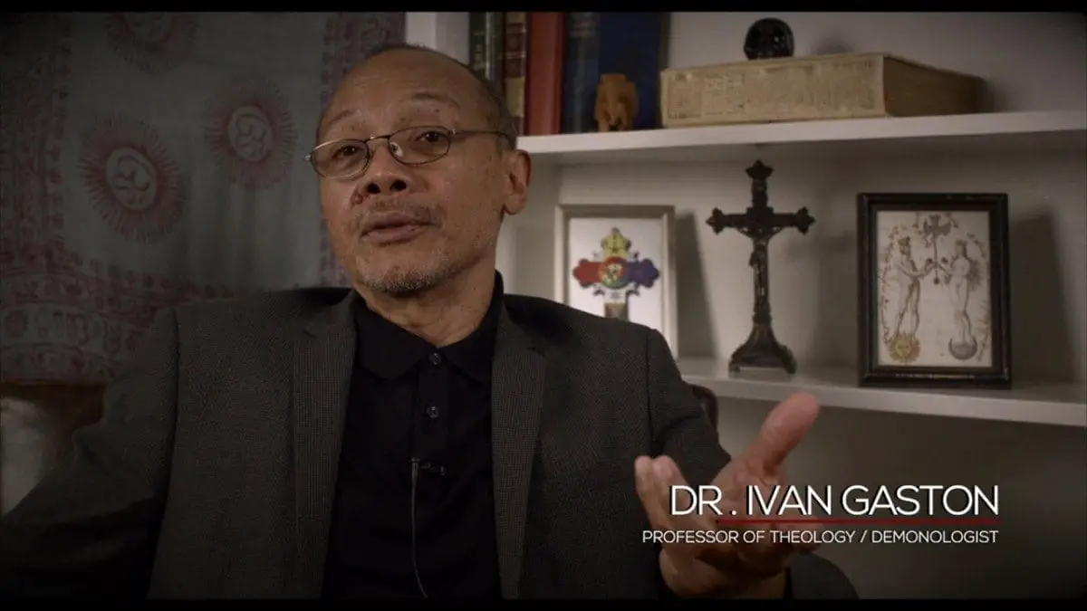 Picture of a middle-aged man sitting in front of a wall of shelves with various occult-themed items on them. Caption states his name is Dr. Ivan Gaston, progessor of theology and demonologist 