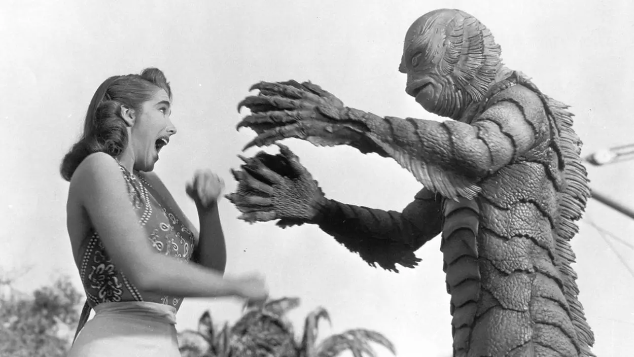Black-and-white still of monstrous Gill Man reaching clawed, webbed fingers out to a screaming white woman.