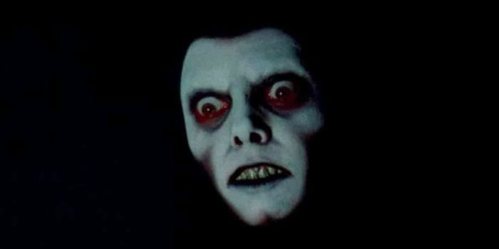 A white face with elaborate black and red makeup around the eyes peers menacingly from the dark.