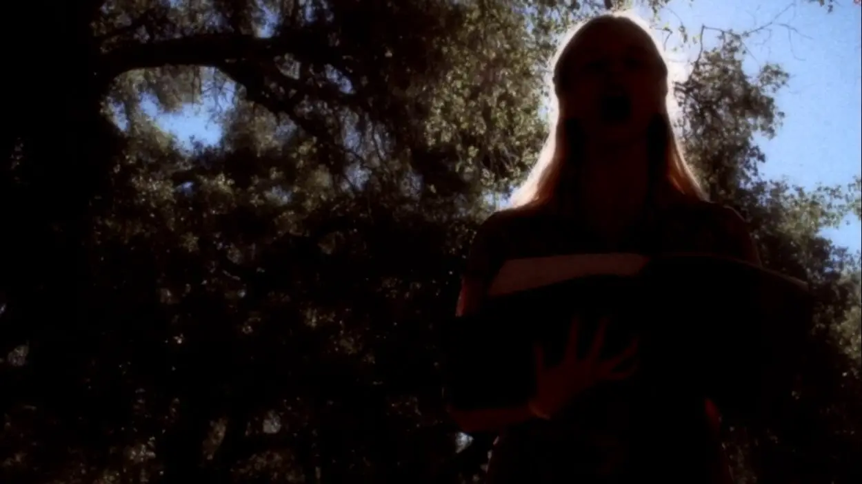 Screenshot of a girl holding a large leather bound book, standing under some trees in the forest. Camera is angled up at her face and she is heavily back lit to the point of silhouette