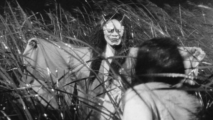 The mother-in-law, in flowing robe, wearing the long-haired, horned, big-eyed demon mask, spreads her arms as she appears from out of the wet, windblown reeds to terrify her daughter-in-law, seen from behind in the foreground.