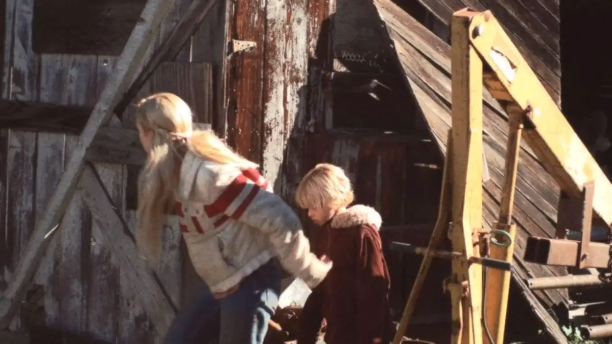 Blond Teenage girl and her young brother peering around the corner of some sort of barn or shed structure with various machinery nearby 
