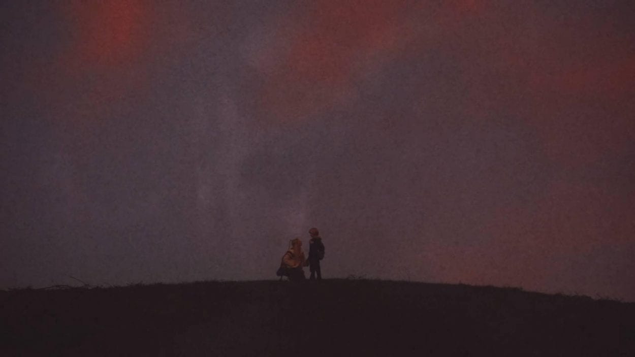 Teenage girl and younger brother standing on a hilltop - silhouetted against the purple dusk sky - the ghostly image of a giant horned devil is visible filling the sky over them