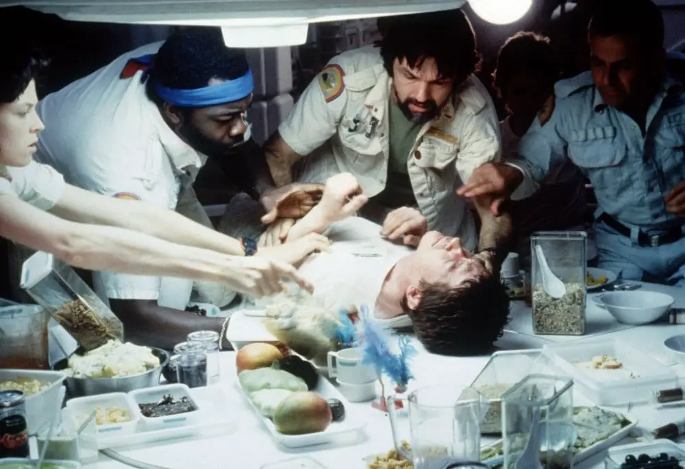 A group of scientists crowd around a sickly man on a table with various specimens and tools on the table in the foreground.