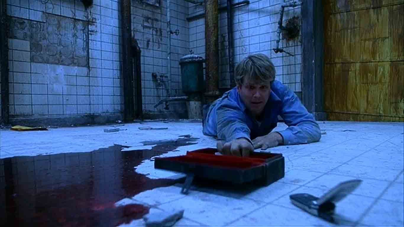 Man desparately reaches for box with saw on bathroom floor next to pool of blood with boarded entrance