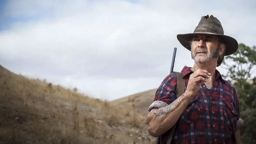 Mick Taylor stands in the wilderness with a rifle slung over his shoulder while he smokes a ciggerate