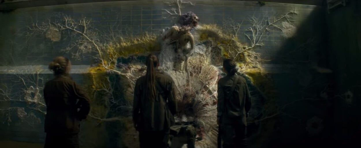 Three characters from Annihilation look on at a body, split at the waste. The top half sits a yard above the bottom half. A ring of moss, flowers and vines circle the body, supporting it.