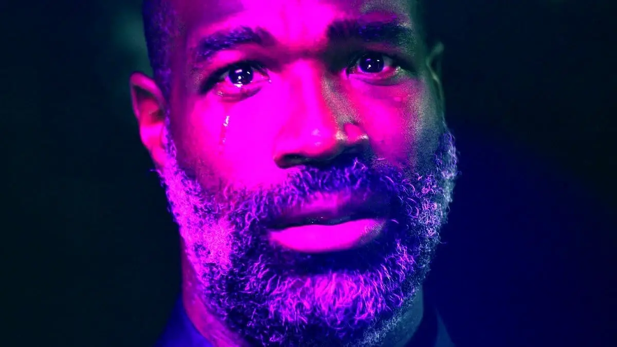 Tunde Adebimpe looking at the camera crying while bathed in a purple otherworldly light