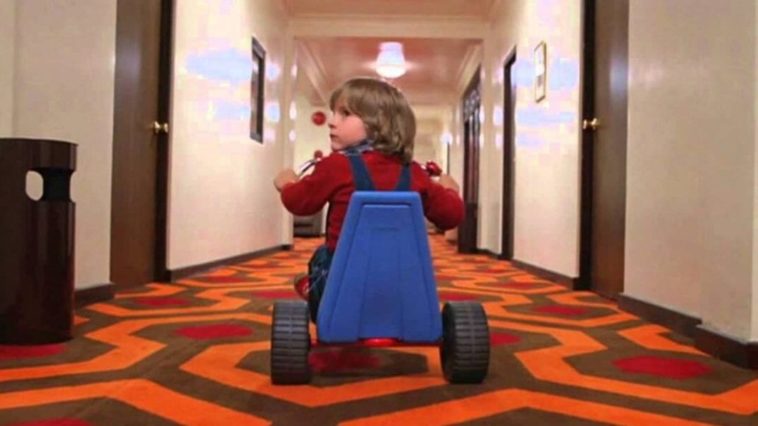Danny Lloyd as Danny Torrance looks over his shoulder uneasily as he pedals down the hotel hallway on his tricycle.