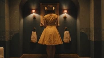 Emma (Christine Kilmer) wearing a 50s style yellow dress, faces a mirror in a bathroom.