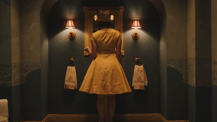 Emma (Christine Kilmer) wearing a 50s style yellow dress, faces a mirror in a bathroom.
