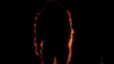 Bigfoot stands in the darkness, highlighted with a dash of light in the distance.