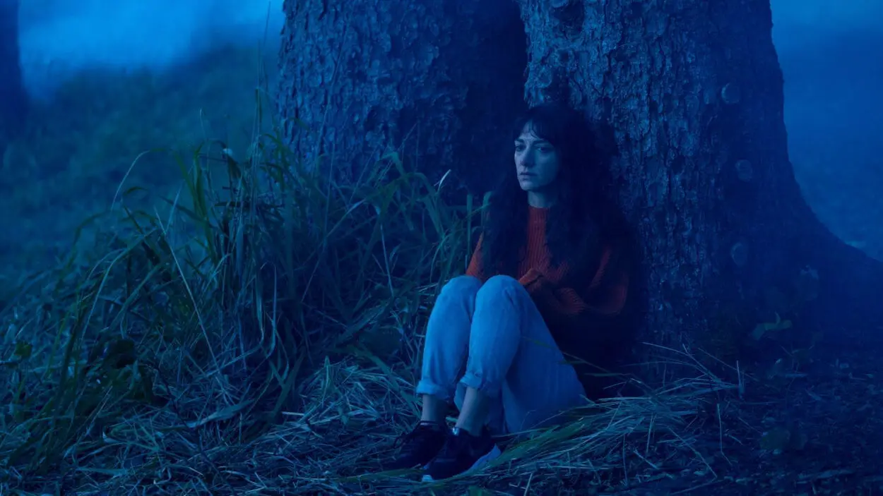 Mina (Sheila Vand) cowering behind a tree in a blue-lit fog-ridden forest.