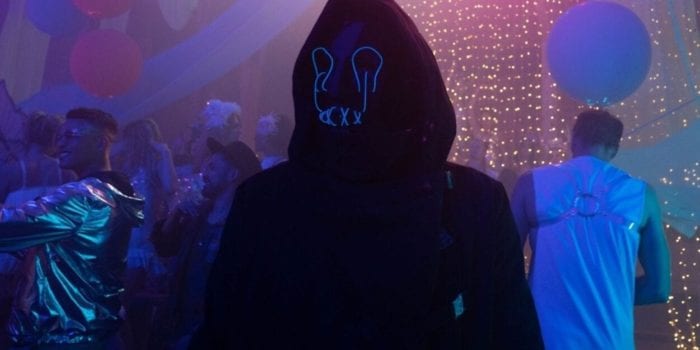 A person in a dark hoodie and glowing blue mask stands in the middle of a party