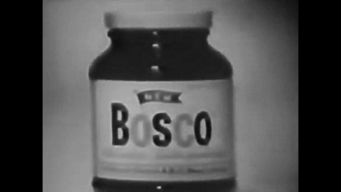 A short, squat, clear glass jar (dark chocolate syrup visible through the glass) with a white lid and white label (Bosco emblazoned across it).