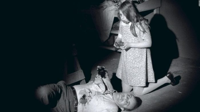 A man, one of his arms missing, lies dead on the floor while, kneeling next to him, a young girl (his daughter), blood smeared around her mouth, holds what appears to be a piece of the man's flesh that she is in the process of eating.