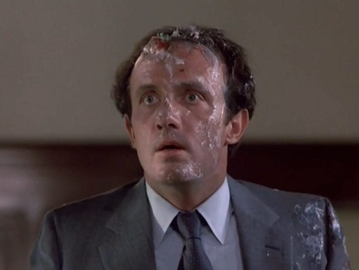 A man, seen from the chest up, wearing a suit and tie, staring wide-eyed, his face covered in food residue.