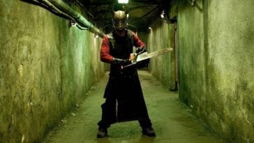 A man wearing a metal mask wields a chainsaw in a dark, dingy hallway.