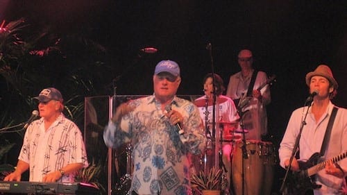 Mike Love, onstage with microphone in hand and other musicians playing in the background.